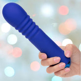 A hand with light pink nails holds the Thicc Chubby Thrusting Wand by CalExotics, a blue ribbed cylindrical silicone toy, against a blurred background with pastel lights.